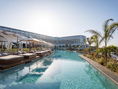 Schöner Hotelpool mit Liegen, Sonnenschirmen, Palmen und modernem Gebäude im Hintergrund.