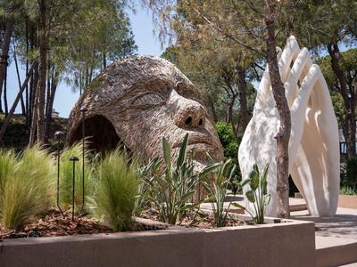 Beeld van een gezicht en grote hand buiten, omringd door planten en bomen.