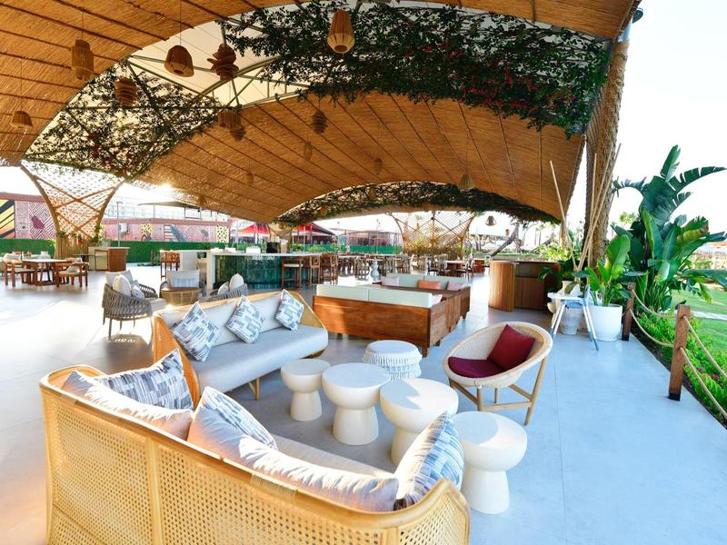 Otevřená odpočinková zóna pod dřevěnou střechou s pohovkami, židlemi a rostlinami vedle zelené venkovní plochy.