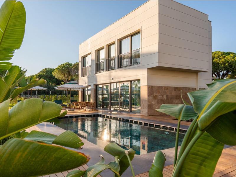 Moderní hotel s velkým bazénem a bujnou tropickou vegetací pod jasnou oblohou.