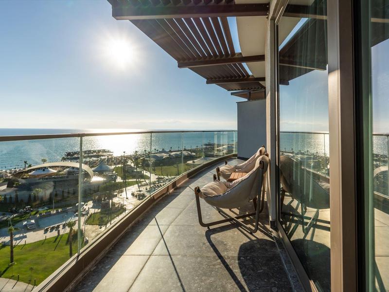 Balkon se dvěma lehátky a výhledem na moře, sluneční světlo odrážející se na skleněné fasádě.
