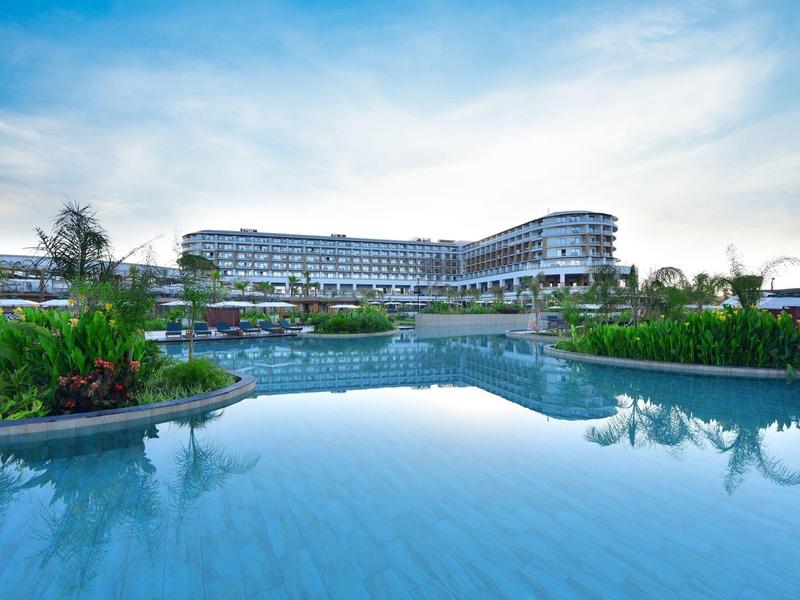 Moderní hotel s velkým bazénem a bujnou zelení pod jasnou oblohou.