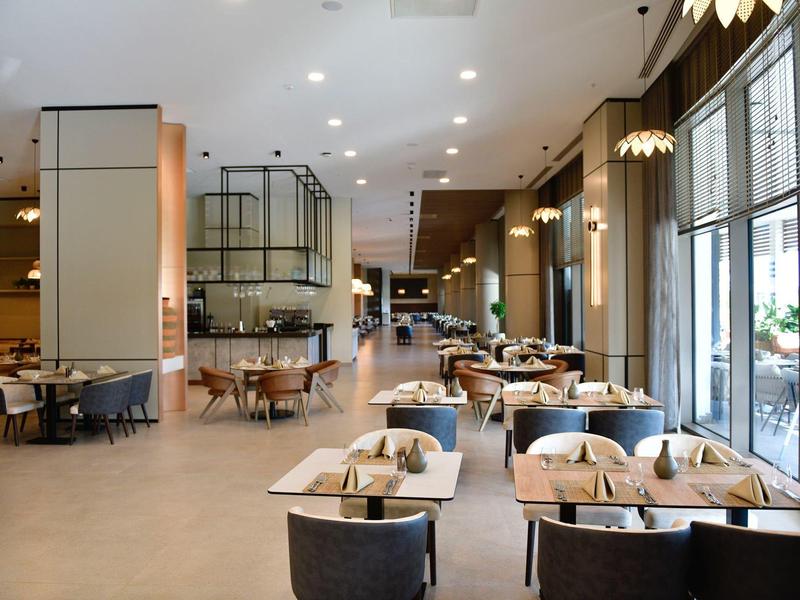 Moderní hotelová restaurace s stoly, židlemi a velkými okny pro denní světlo.