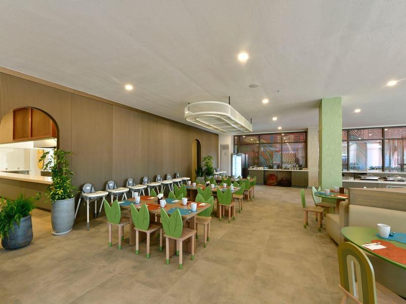 Moderní jídelna s dlouhými stoly, zelenými židlemi a velkými okny vpouštějícími spoustu přirozeného světla.