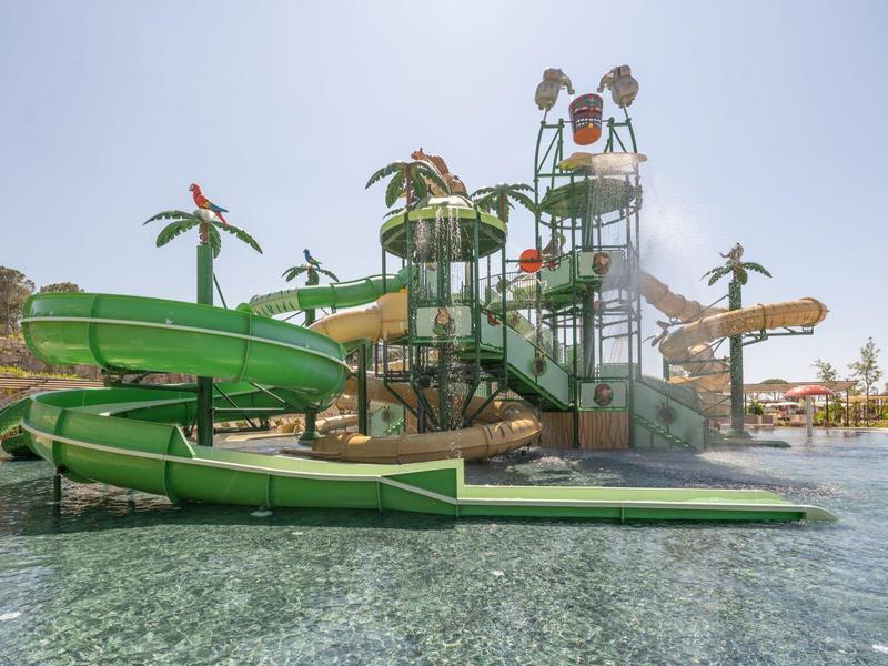 Vodní park se zelenými tobogány, palmami a bazény pod jasnou oblohou.