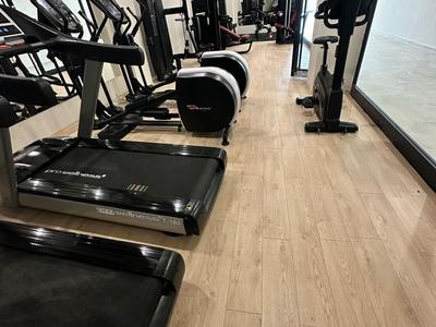 Světelná posilovna s moderními běžeckými pásy, eliptickými stroji a závažími na dřevěné podlaze.