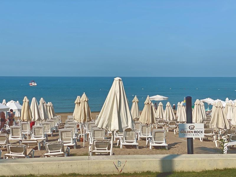 Přeplážovaný písečný pláž s lehátky a bílými slunečníky, klidné moře v pozadí.