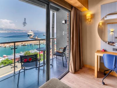Hotel room with balcony view of harbor, chairs and table outside, desk with mirror inside.