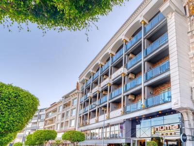 Multi-story hotel with balconies and green trees in front on a sunny street.