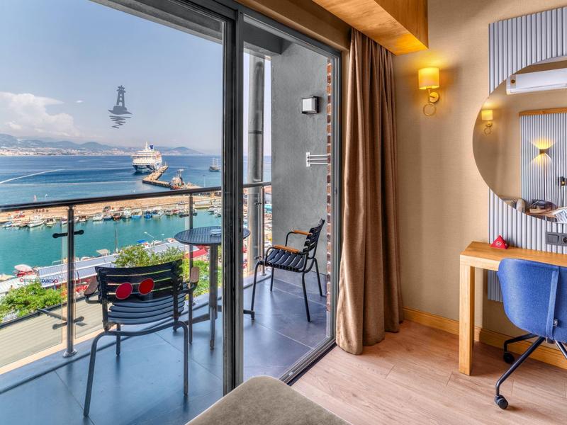 Hotel room with balcony view of harbor, chairs and table outside, desk with mirror inside.