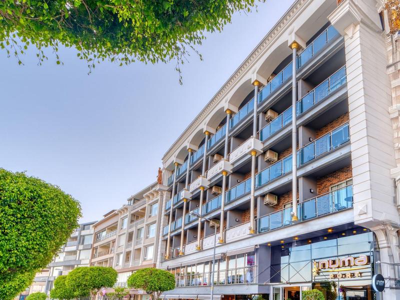 Multi-story hotel with balconies and green trees in front on a sunny street.
