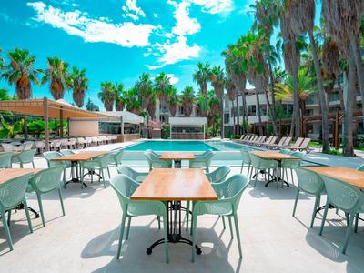 Buiten eetgedeelte hotel met tafels, stoelen, palmbomen en een zwembad onder een blauwe lucht.