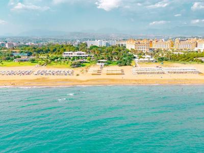 Een resort aan het strand met rijen ligbedden, groene tuinen en een helder blauwe zee onder een deels bewolkte hemel.