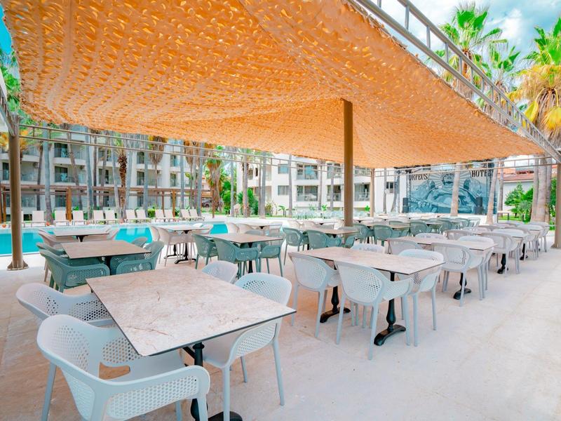 Buitenterras van hotel met tafels, stoelen en schaduwdoek bij een zwembad.