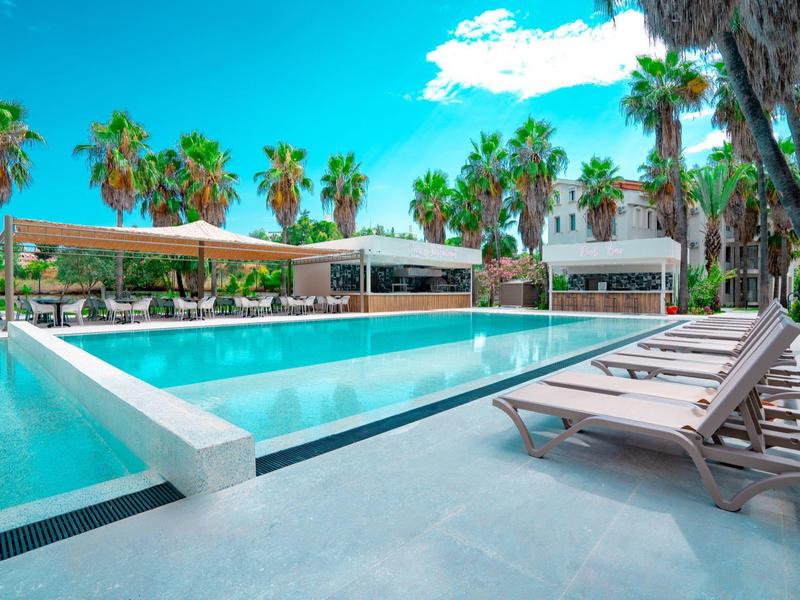 Buitenzwembad met ligstoelen, omringd door palmbomen en moderne gebouwen onder een blauwe hemel.