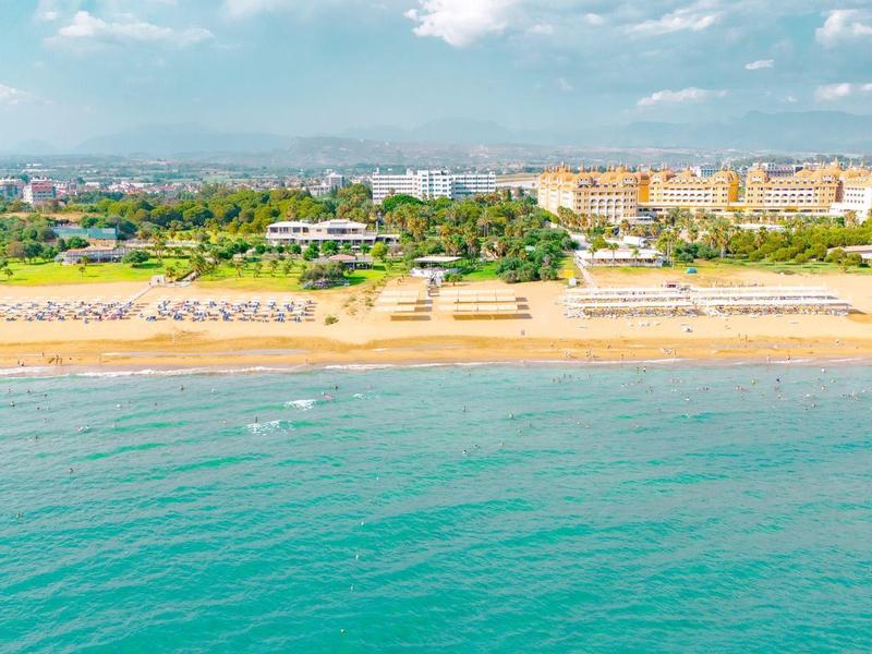 Een resort aan het strand met rijen ligbedden, groene tuinen en een helder blauwe zee onder een deels bewolkte hemel.