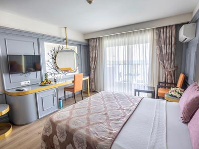 Habitación de hotel luminosa con cama grande, escritorio, TV y balcón con cortinas.