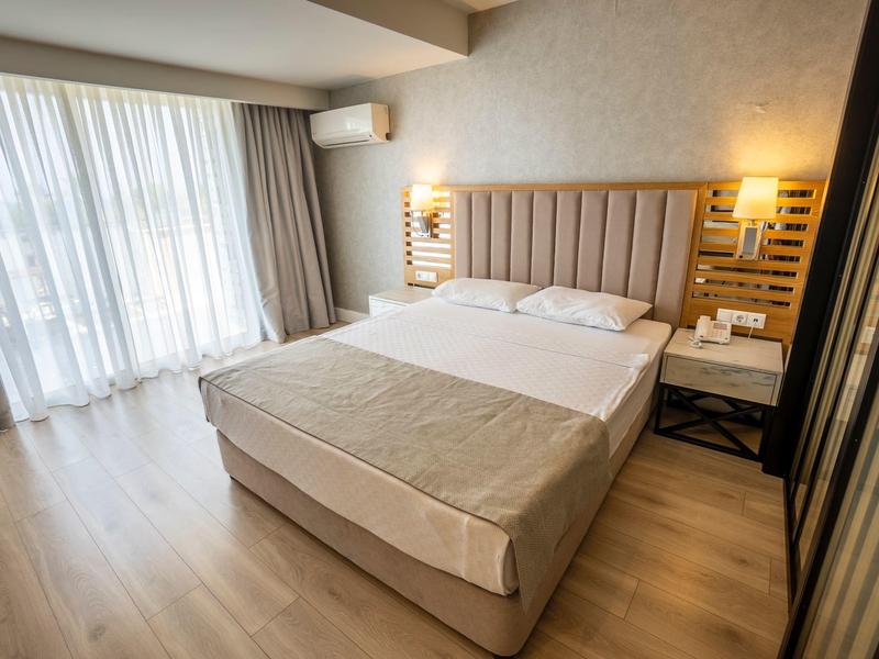 Bright hotel room with large bed, soft lighting, wooden floors, and sheer curtains.