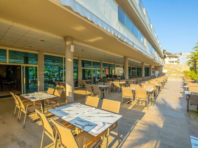 Outdoor hotel dining area with tables and chairs under a covered walkway in sunny weather.