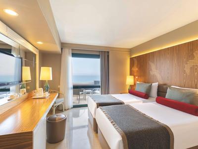 Modern hotelkamer met twee bedden, bureau en uitzicht op balkon en zee.