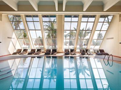 Piscine intérieure avec grandes fenêtres et chaises longues face aux palmiers.