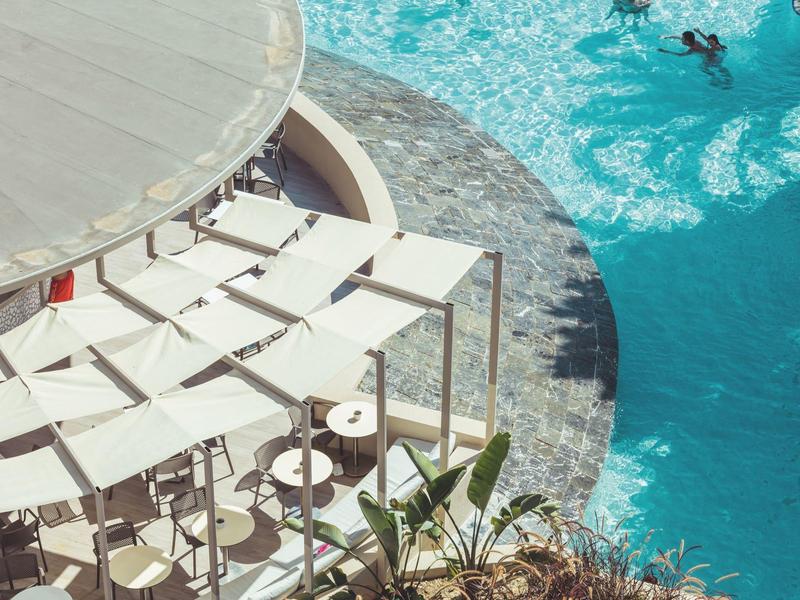 Piscine bleue avec des nageurs et une zone de repos avec chaises blanches et tables rondes.