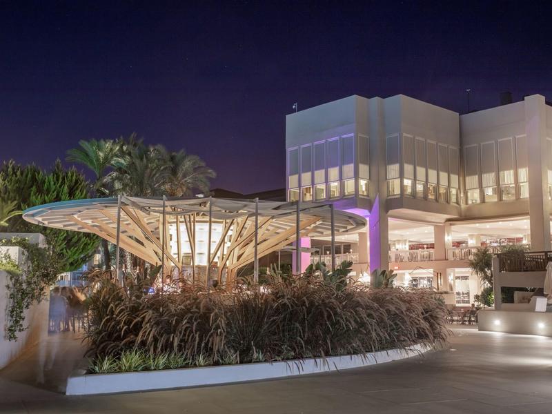Hôtel moderne éclairé la nuit avec une terrasse et des plantes décoratives.
