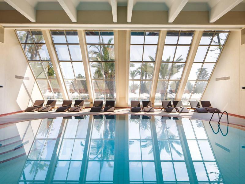 Piscine intérieure avec grandes fenêtres et chaises longues face aux palmiers.