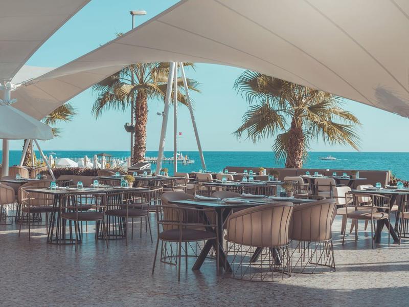 Terrasse d'hôtel en bord de mer avec parasols, tables et chaises sous un ciel clair.