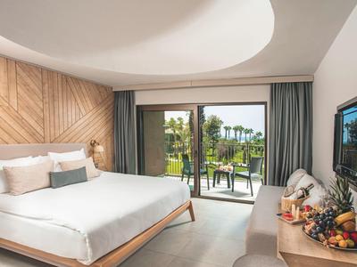 Světý hotelový pokoj s manželskou postelí, balkonem a výhledem na zahradu s palmami.