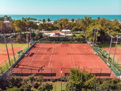 Dwa czerwone korty tenisowe otoczone zielonymi ogrodzeniami z drzewami i morzem w tle