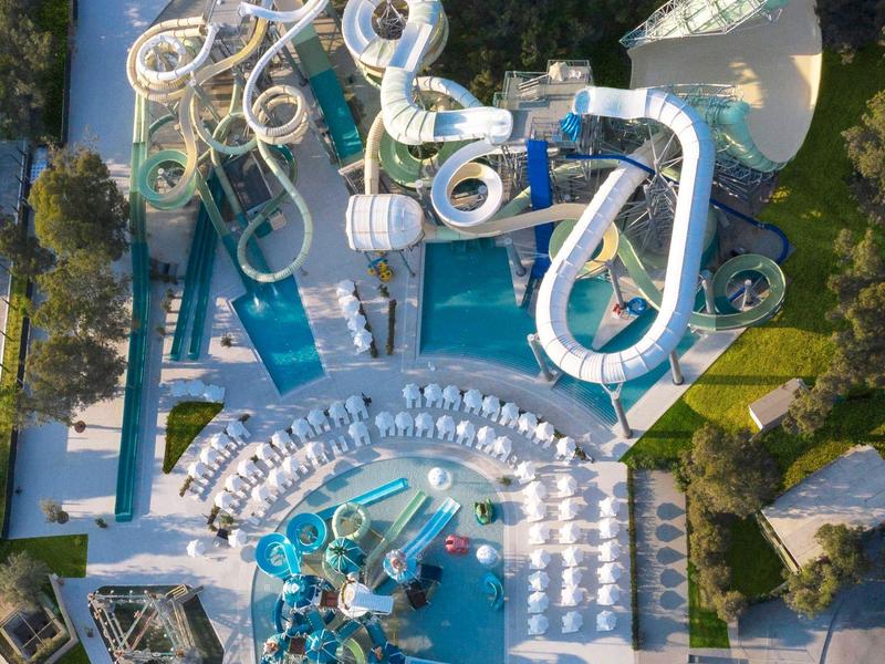 Pohled z ptačí perspektivy na vodní park s několika skluzavkami a bazény, obklopený lehátky a zelení.