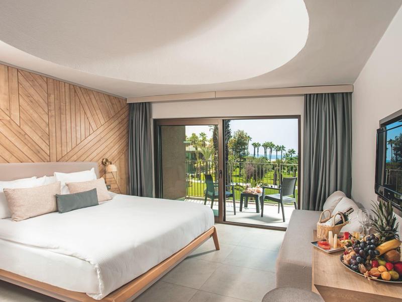 Světý hotelový pokoj s manželskou postelí, balkonem a výhledem na zahradu s palmami.