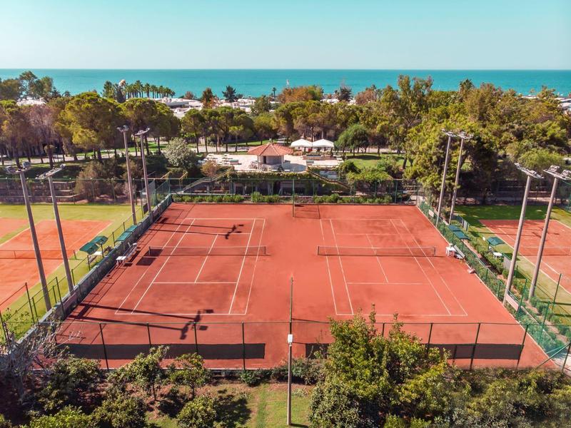 Červené tenisové kurty obklopené zelení s výhledem na oceán.