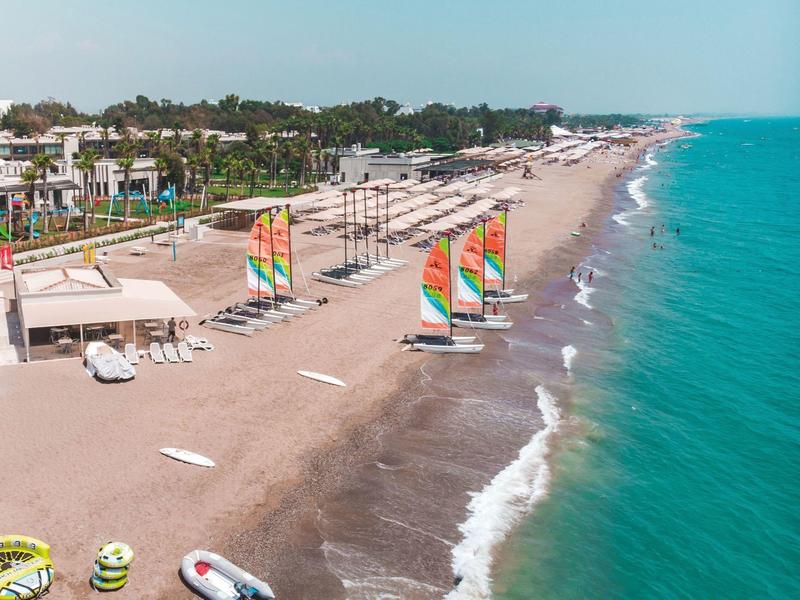 Pohled na klidnou pláž s barevnými plachetními loděmi, slunečnímiky a modrým mořem.