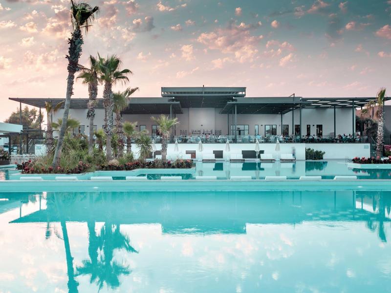 Moderní hotelový bazén se slunečními lehátky a palmami při západu slunce.