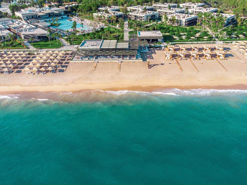 Letecký pohled na resort s písečnou pláží, slunečníky a tyrkysovým mořem.