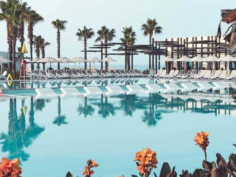 Velký hotelový bazén s lehátky, palmami a kvetoucími rostlinami v popředí pod jasnou oblohou.