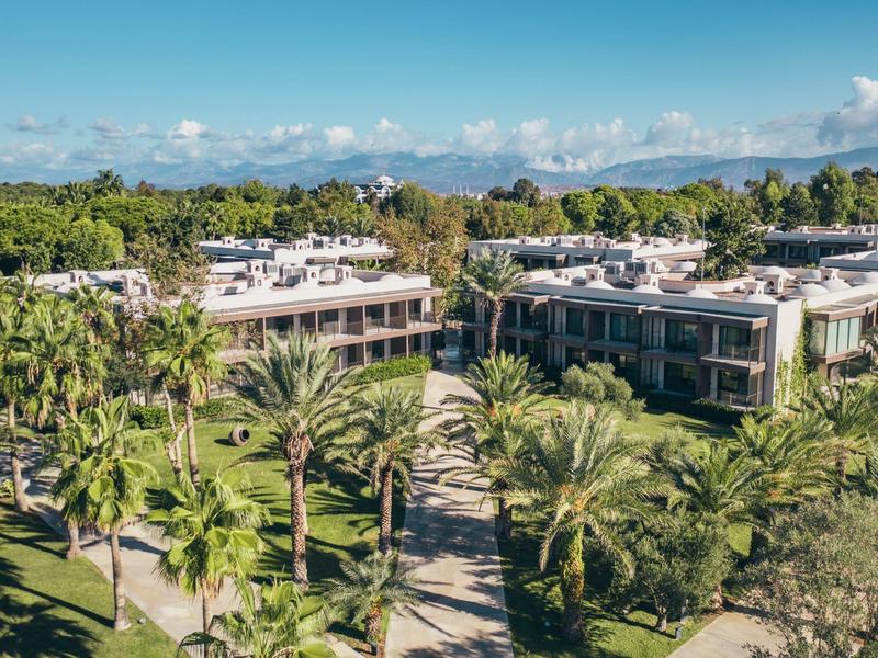 Moderní hotel s plochými střechami obklopený zahradami a palmami pod modrou oblohou.