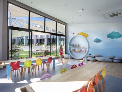 Heller Kinderraum mit bunten Stühlen, großen Fenstern, Wanddekorationen und gemütlichen Sitzkissen.