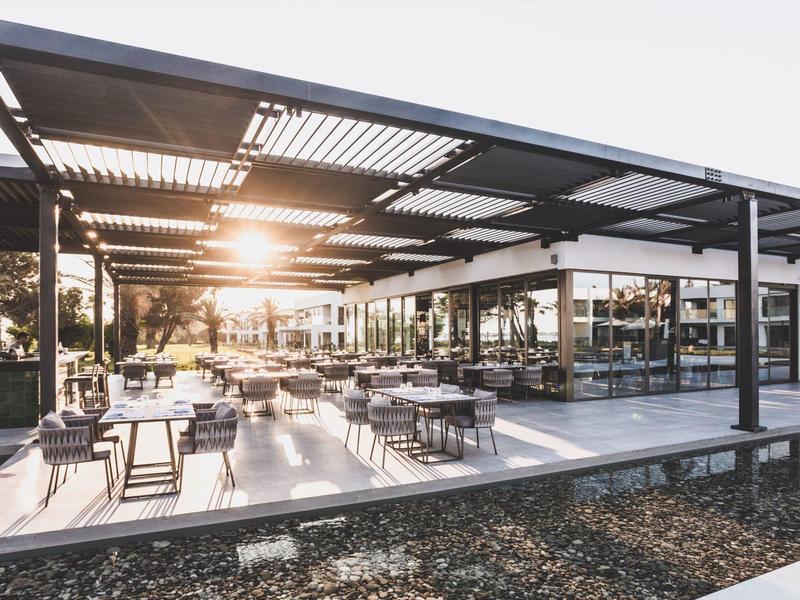 Modernes, offenes Restaurant mit vielen Tischen unter einem großen, schattenspendenden Dach bei Sonnenuntergang.