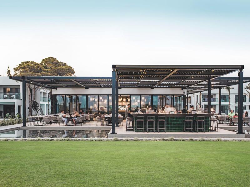 Modernes, offenes Restaurant mit großem Außensitzbereich, Holz- und Metallstruktur, grünem Rasen im Vordergrund.