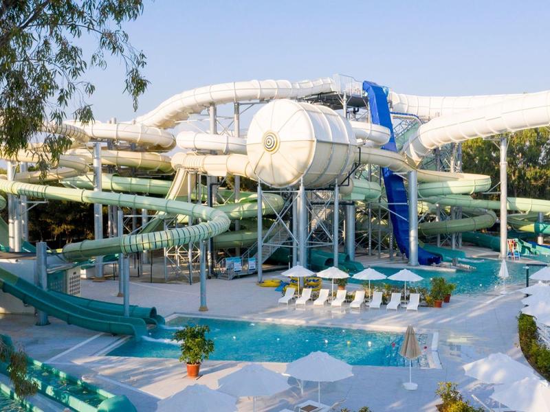 Großes Schwimmbecken mit Wasserrutschen und Sonnenschirmen in einem Freizeitpark bei Tageslicht.