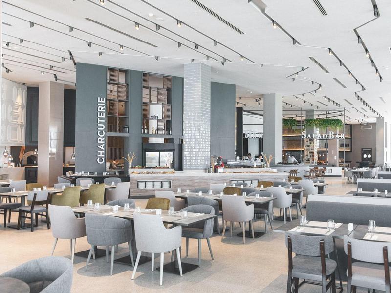 Moderne, helle Cafeteria mit vielen Tischen, grauen Stühlen und einer Kuchentheke im Hintergrund.