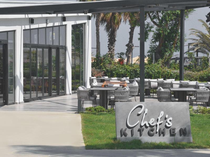 Moderner Außenbereich eines Restaurants mit Glasfenstern, Tischen, Palmen und einem Schild "Chef's Kitchen".