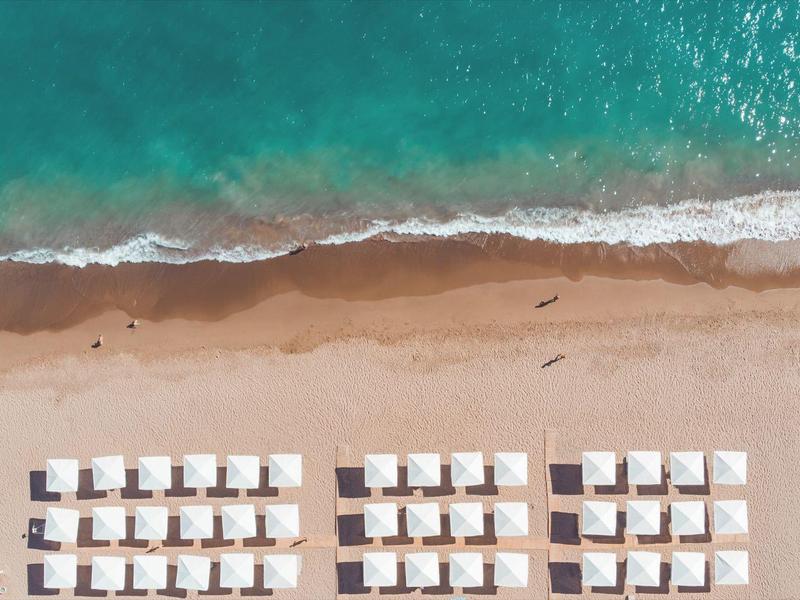 Drohnenansicht von Strand mit Reihen weißer Sonnenliegen und türkisfarbenem Meer im Hintergrund.