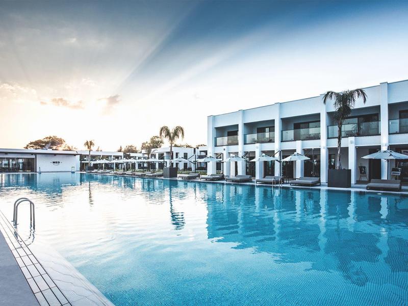 Großer Außenpool vor modernem weißen Gebäude mit Palmen und sanftem Sonnenlicht am Himmel.