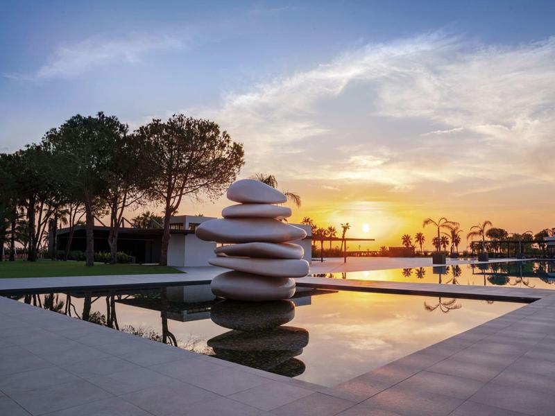 Sonniger Himmel über ruhigem Pool mit großer Steinskulptur und Bäumen am Rand.