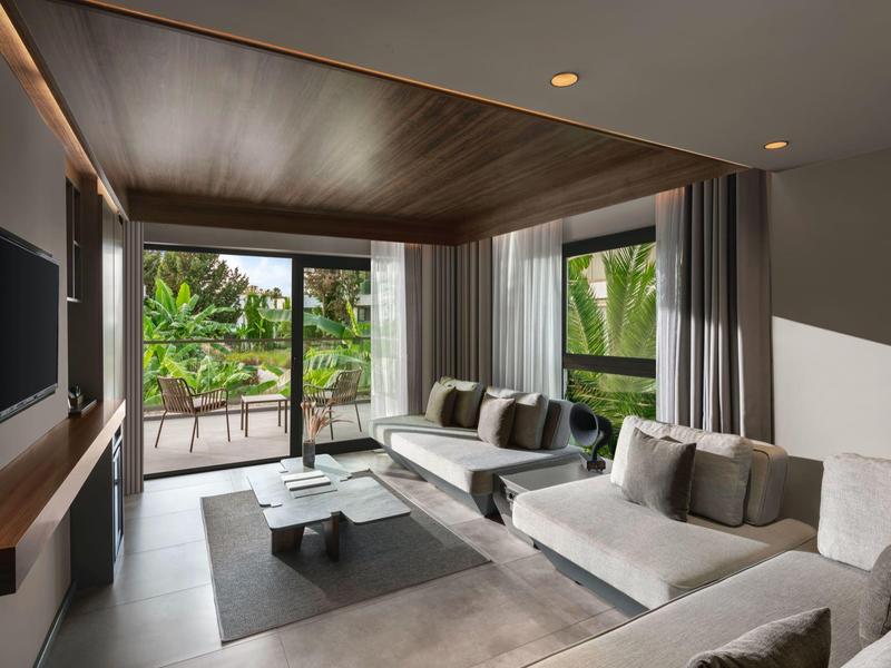 Modernes Wohnzimmer mit großen Fenstern, grauen Sofas, Holzboden und einem TV an der Wand.