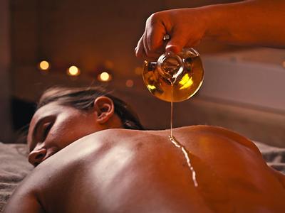 Frau entspannt auf Massagetisch, Öl wird sanft auf ihren Rücken gegossen, warme Atmosphäre.